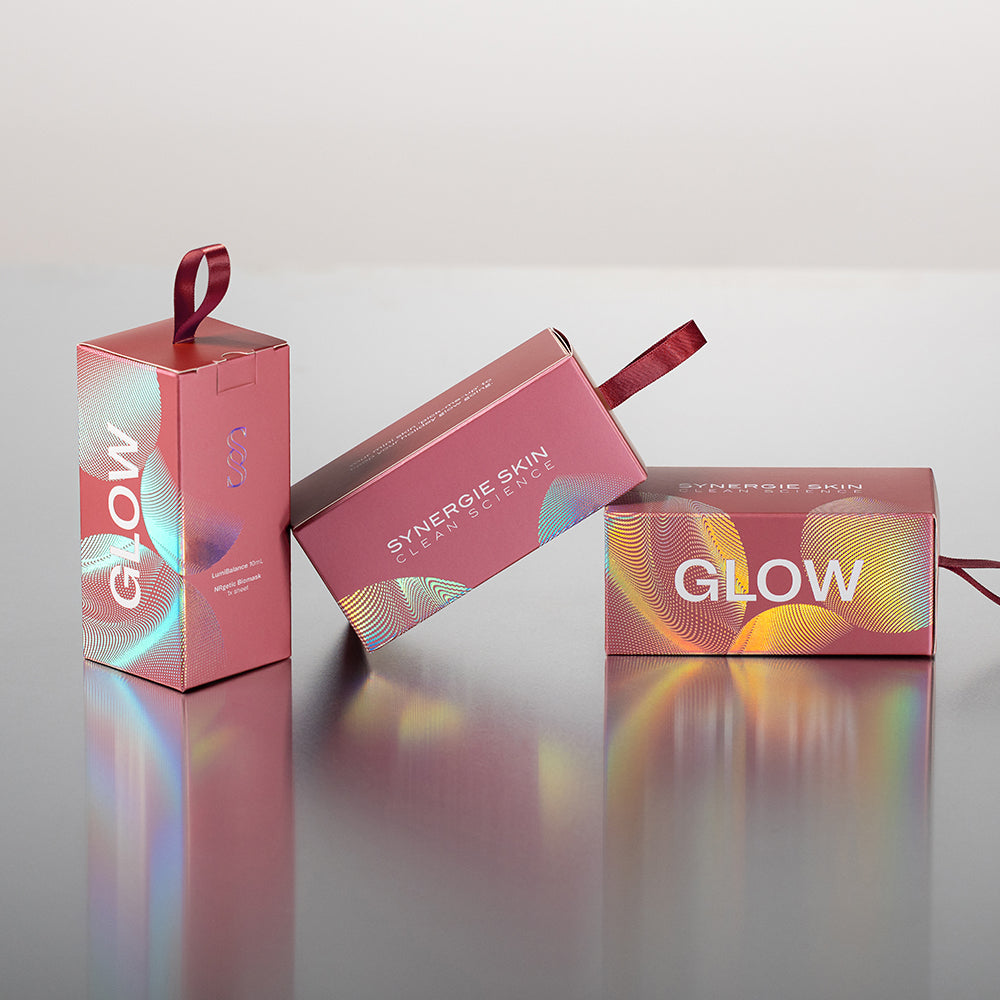 Three pink cosmetic boxes with iridescent holographic accents and red ribbon handles, arranged on a reflective surface against a light gray background. Visible text includes GLOW and SYNERGIE SKIN.