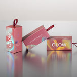 Three pink cosmetic boxes with iridescent holographic accents and red ribbon handles, arranged on a reflective surface against a light gray background. Visible text includes GLOW and SYNERGIE SKIN.